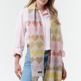 Woman modelling a multi coloured heart printed scarf, with fringe, with a pink blouse and blue jeans photographed on a light background.