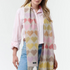 Woman modelling a multi coloured heart printed scarf, with fringe, with a pink blouse and blue jeans photographed on a light background.