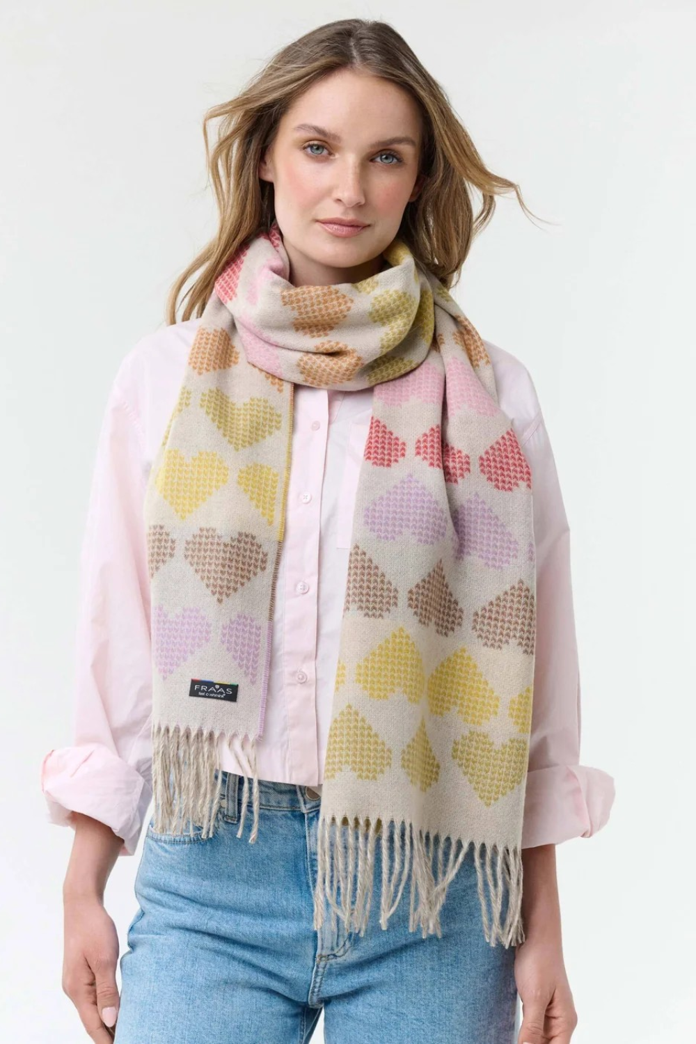 Woman modelling a multi coloured heart printed scarf, with fringe, with a pink blouse and blue jeans photographed on a light background.