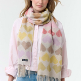 Woman modelling a multi coloured heart printed scarf, with fringe, with a pink blouse and blue jeans photographed on a light background.