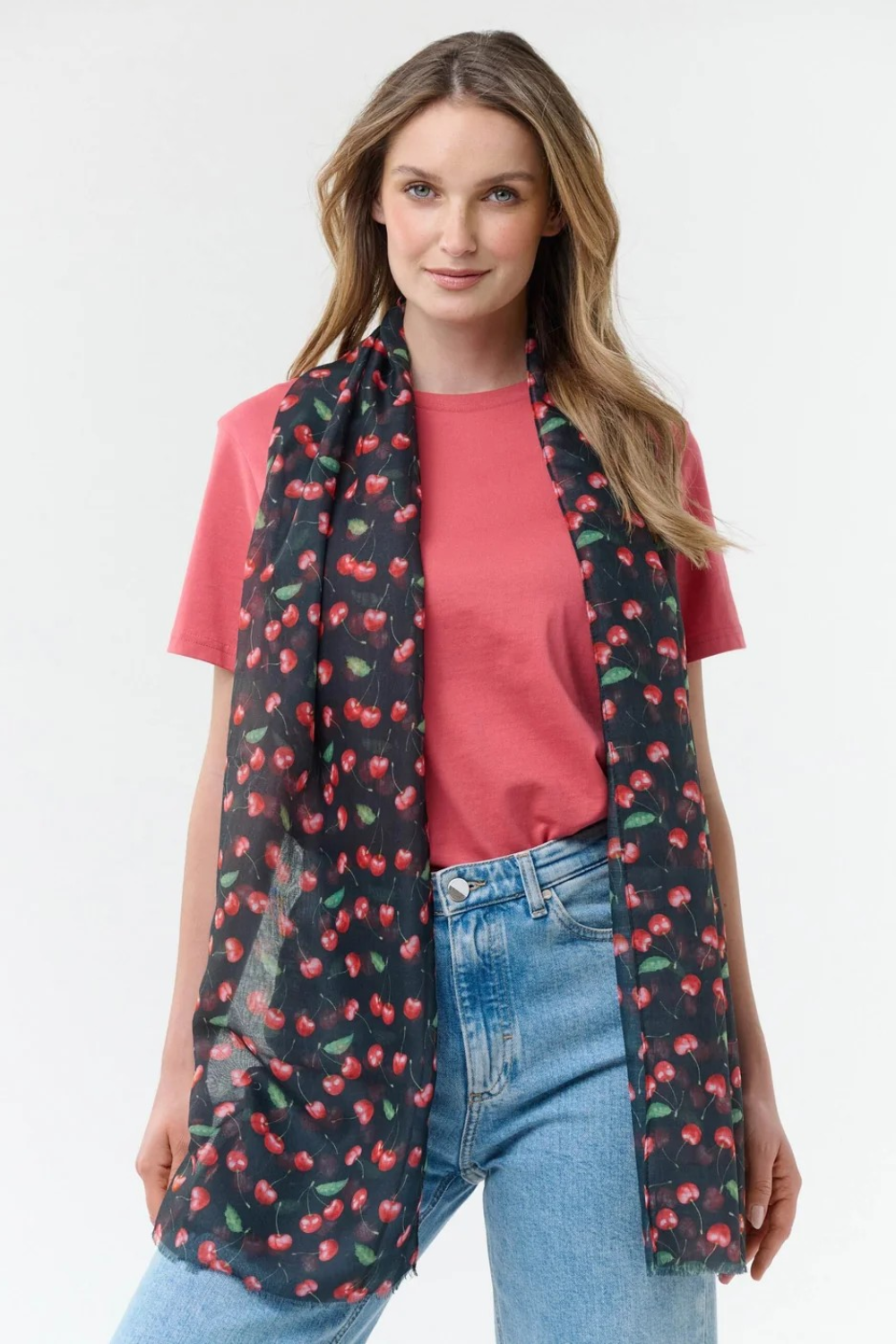 Woman modelling a black base scarf with a cherry print  on a red t-shirt with blue jeans on a light background.