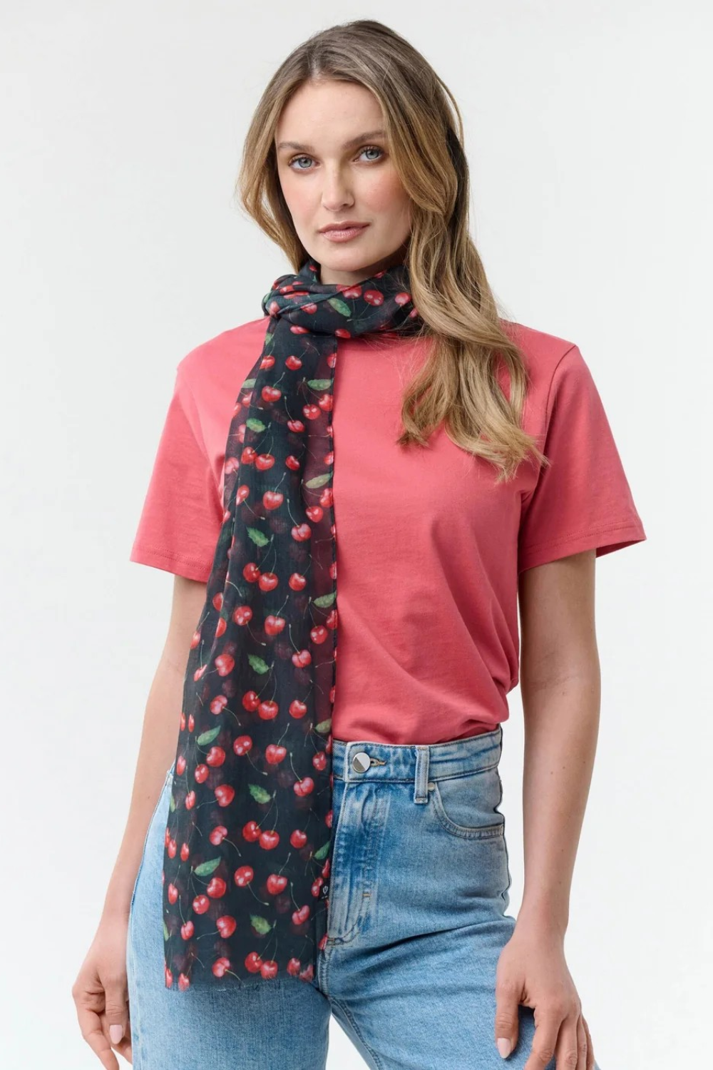 Woman modelling a black base scarf with a cherry print  on a red t-shirt with blue jeans on a light background.