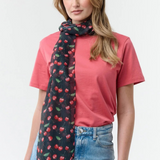 Woman modelling a black base scarf with a cherry print  on a red t-shirt with blue jeans on a light background.