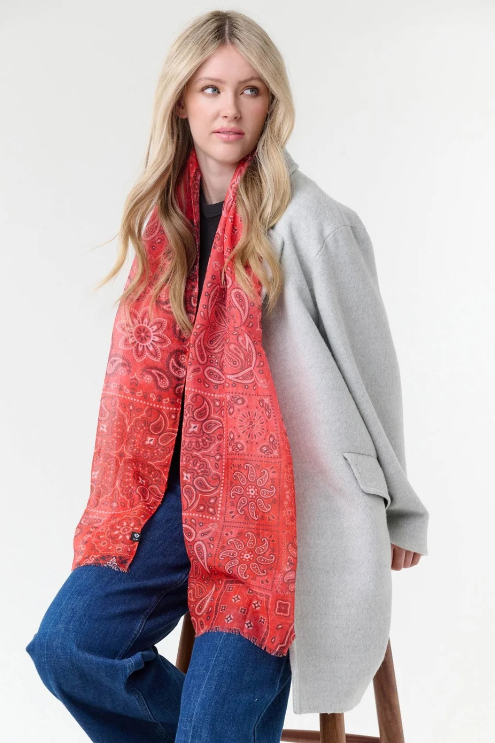 Woman modelling a red paisley scarf with a black top, blue jeans, and a grey coat, photographed on a light background.