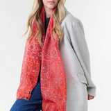 Woman modelling a red paisley scarf with a black top, blue jeans, and a grey coat, photographed on a light background.