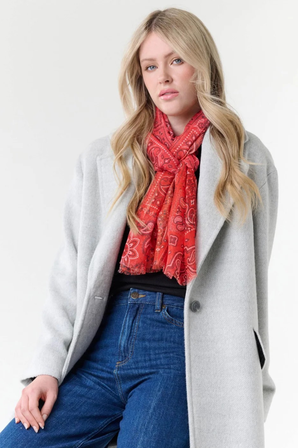 Woman modelling a red paisley scarf with a black top, blue jeans, and a grey coat, photographed on a light background.