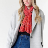 Woman modelling a red paisley scarf with a black top, blue jeans, and a grey coat, photographed on a light background.