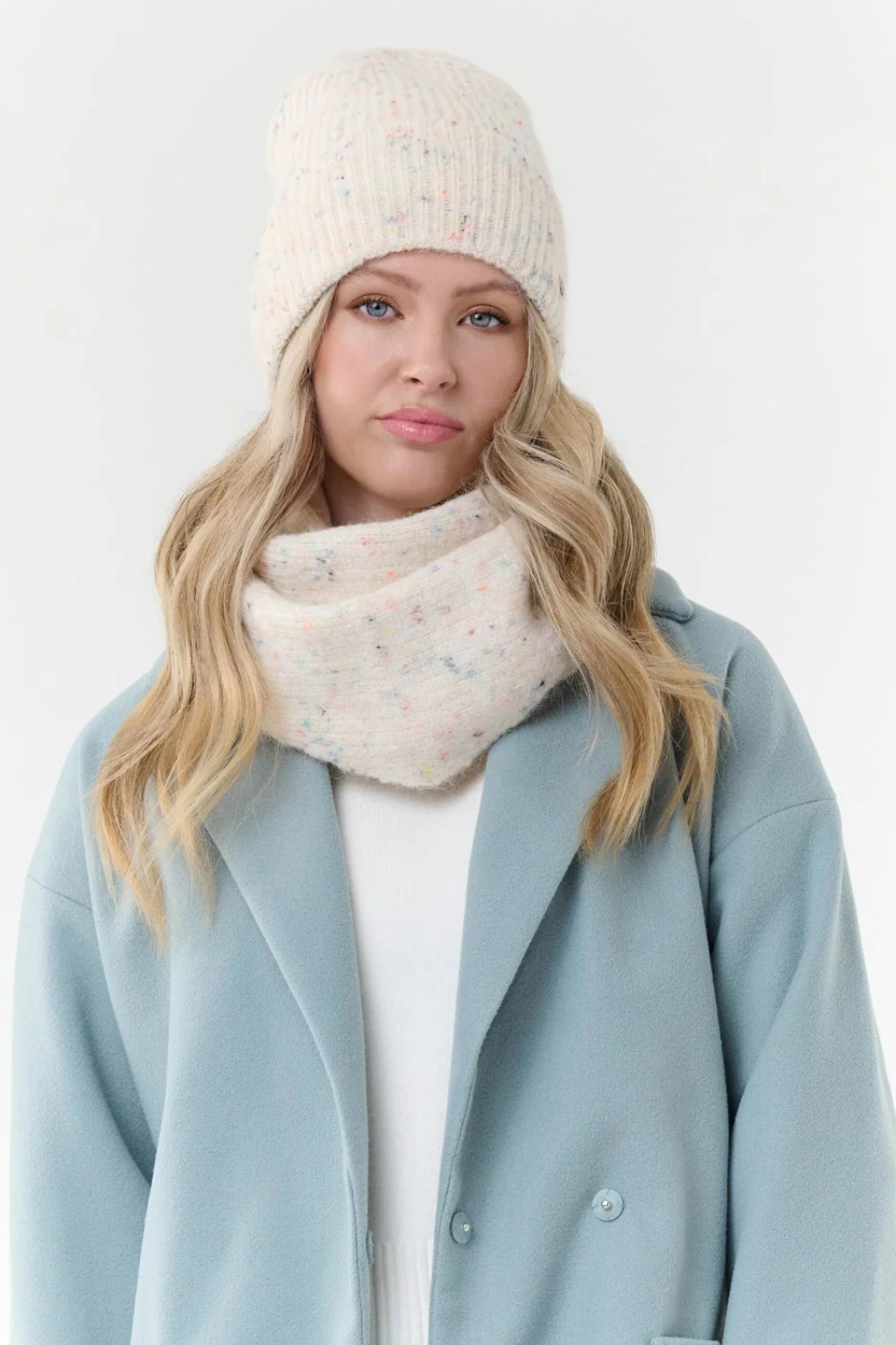 Woman modelling a cream toque with multi coloured flecks, with a matching scarf, a cream sweater and blue coat  photographed on a light background.