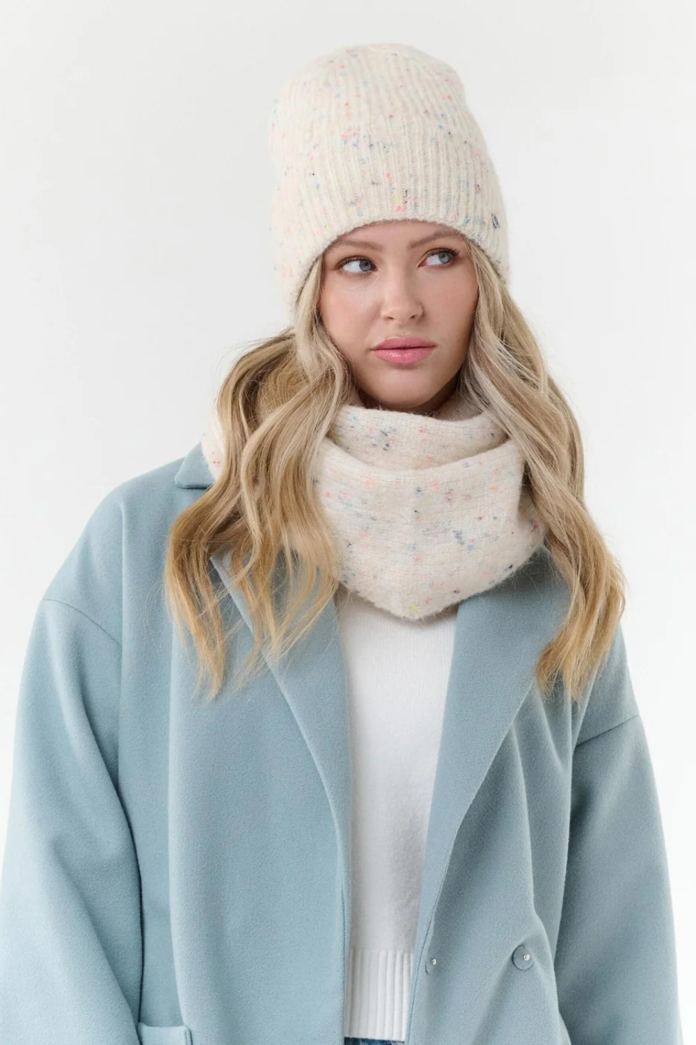 Woman modelling a cream scarf with multi coloured flecks, with a matching toque, cream sweater and blue coat, photographed on a light background.
