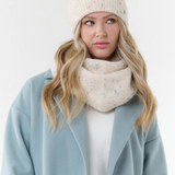 Woman modelling a cream scarf with multi coloured flecks, with a matching toque, cream sweater and blue coat, photographed on a light background.