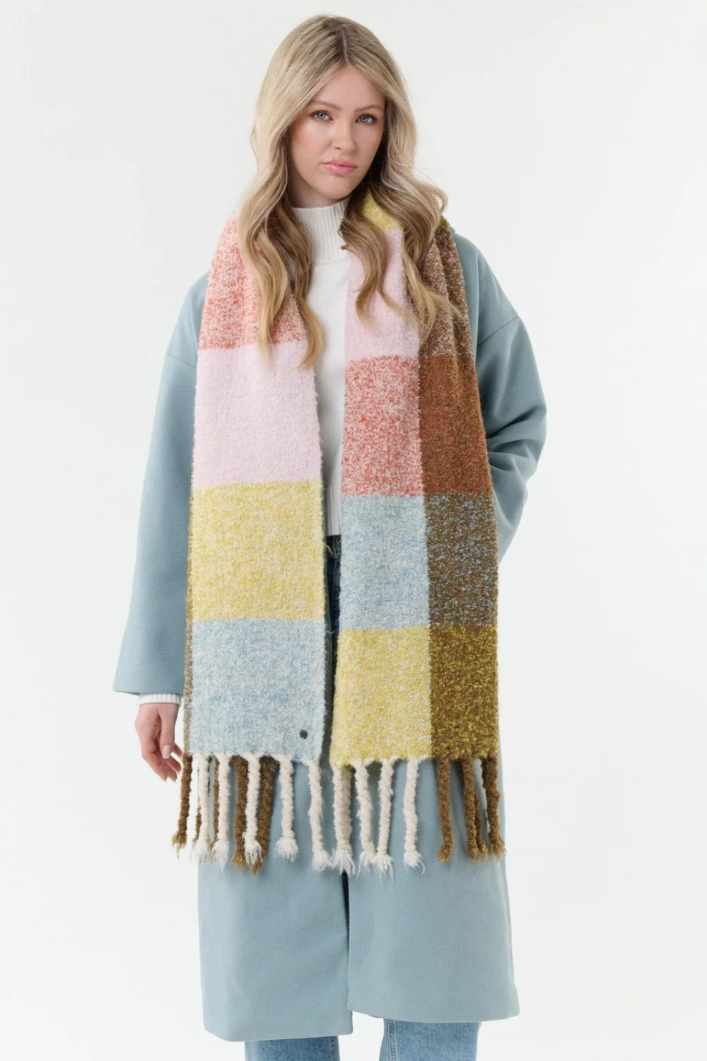 Woman modelling a multi coloured checkered scarf, with fringe, with a white t shirt, blue jeans and a blue coat on a light background.