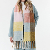 Woman modelling a multi coloured checkered scarf, with fringe, with a white t shirt, blue jeans and a blue coat on a light background.