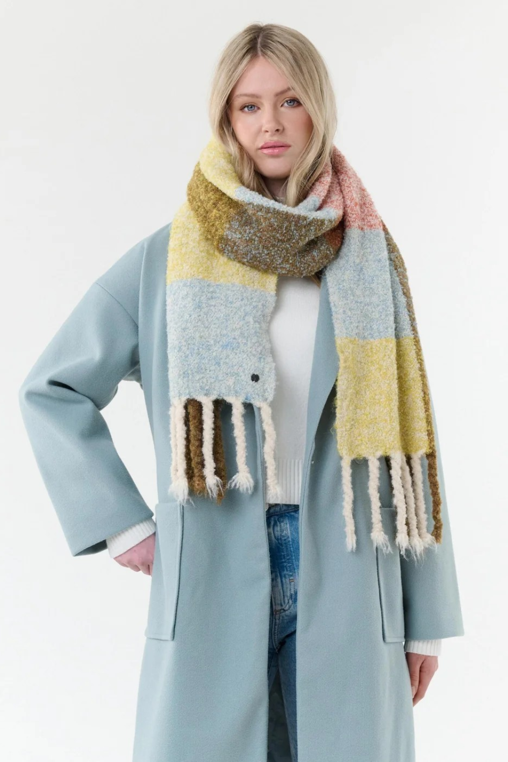 Woman modelling a multi coloured checkered scarf, with fringe, with a white t shirt, blue jeans and a blue coat on a light background.