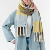 Woman modelling a multi coloured checkered scarf, with fringe, with a white t shirt, blue jeans and a blue coat on a light background.