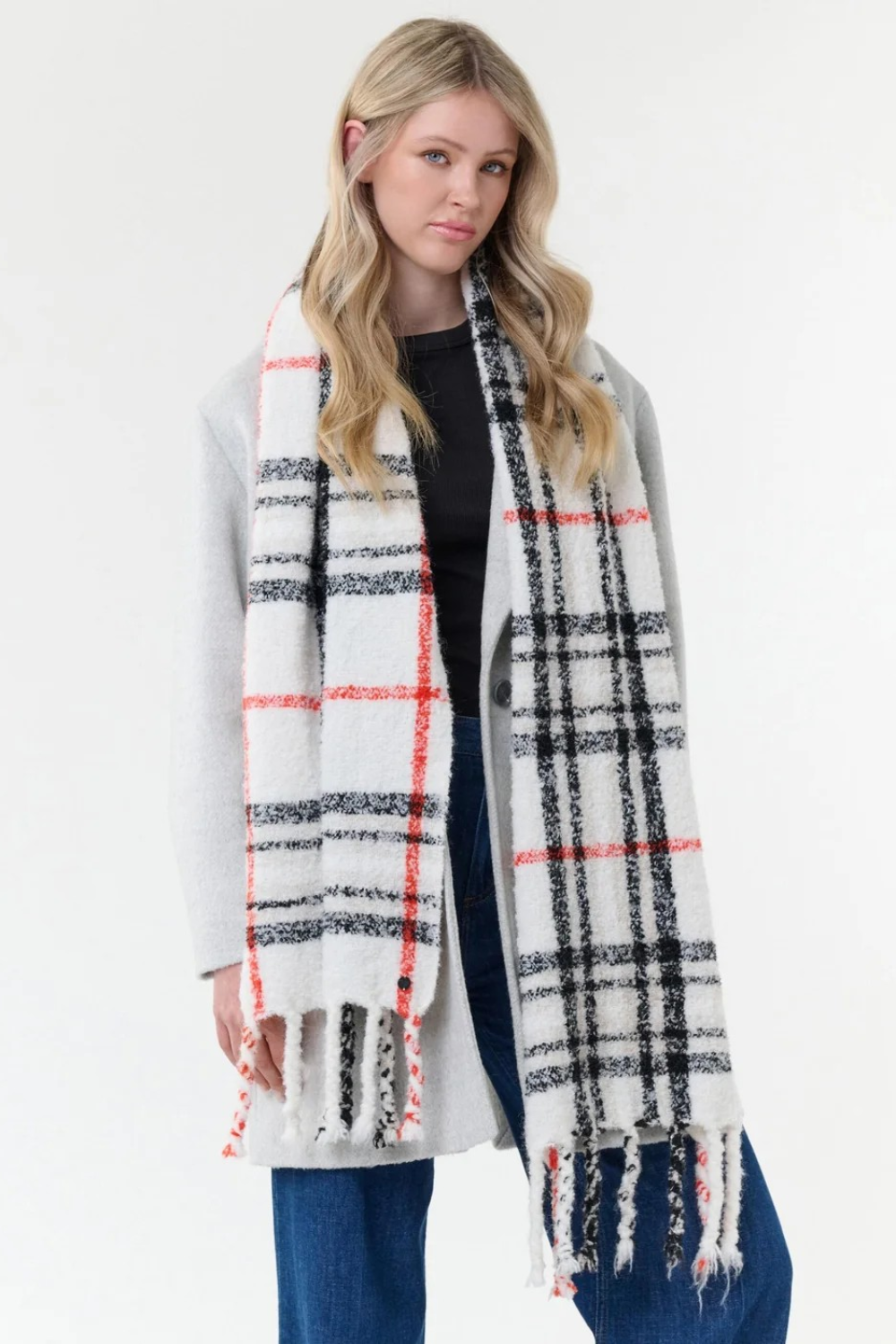 Woman modelling a cream, black and orange plaid scarf with fringe with a black shirt, blue jeans and a grey coat on a light background.
