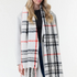 Woman modelling a cream, black and orange plaid scarf with fringe with a black shirt, blue jeans and a grey coat on a light background.