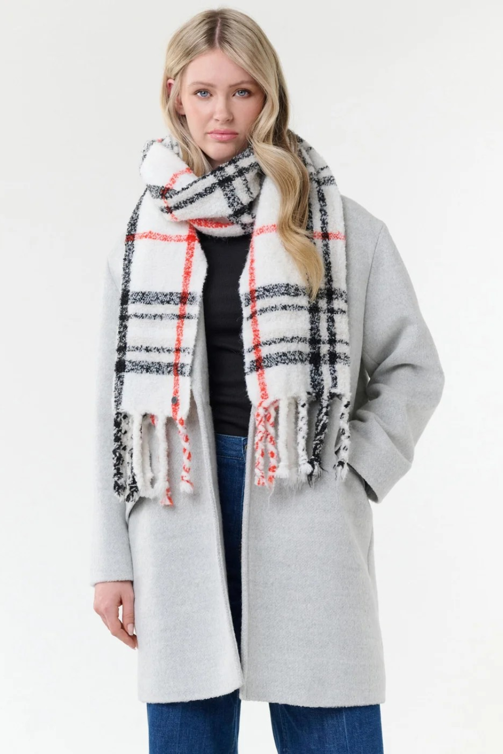 Woman modelling a cream, black and orange plaid scarf with fringe with a black shirt, blue jeans and a grey coat on a light background.