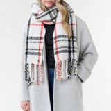 Woman modelling a cream, black and orange plaid scarf with fringe with a black shirt, blue jeans and a grey coat on a light background.