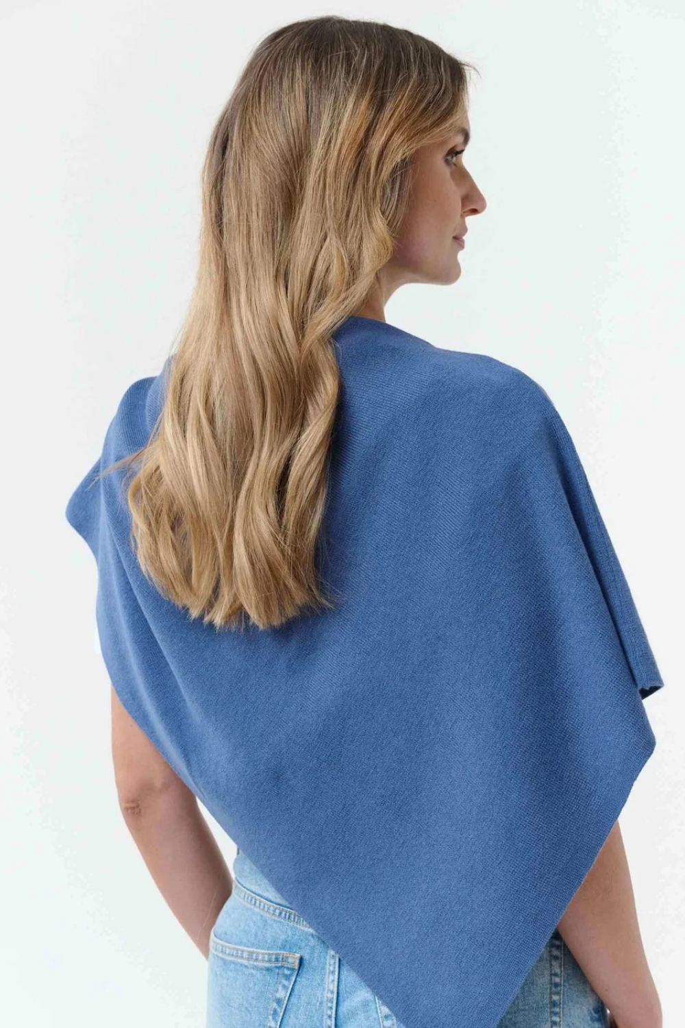 Woman, shown from behind, modelling a blue poncho, with blue jeans, against light background.