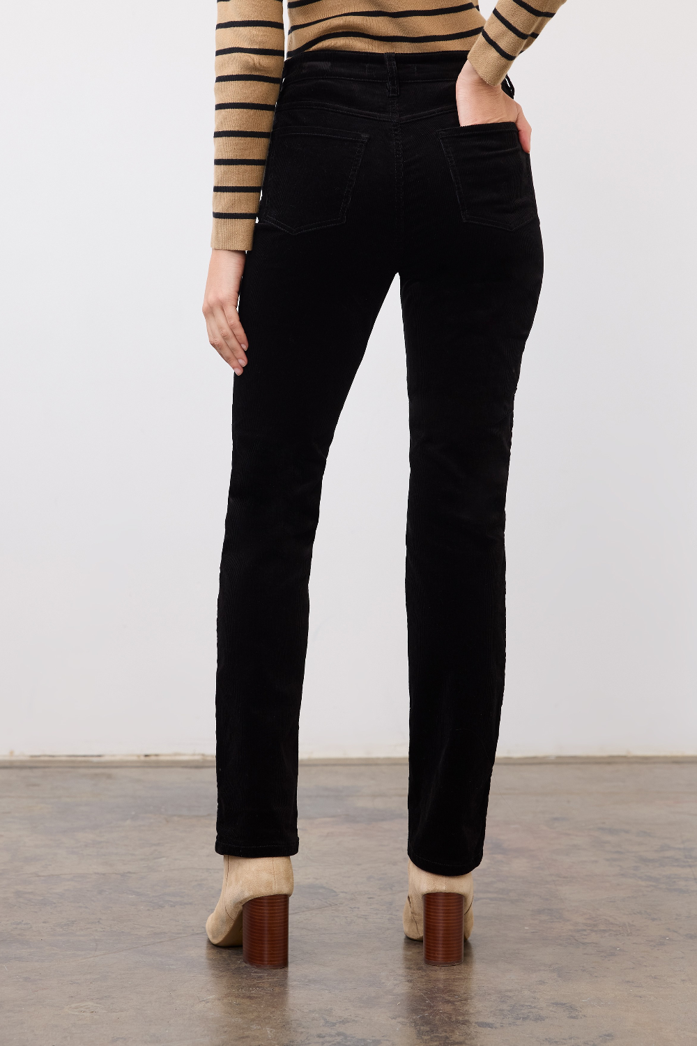 Woman, shown from behind, modelling a straight leg black corduroy pant with a tan and black striped sweater and tan boots photographed against a light background.