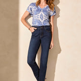 Woman wearing a blue patterned top and dark jeans against a beige wall.