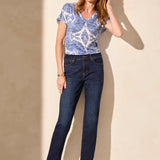 Woman wearing a blue patterned shirt and dark jeans against a beige wall.