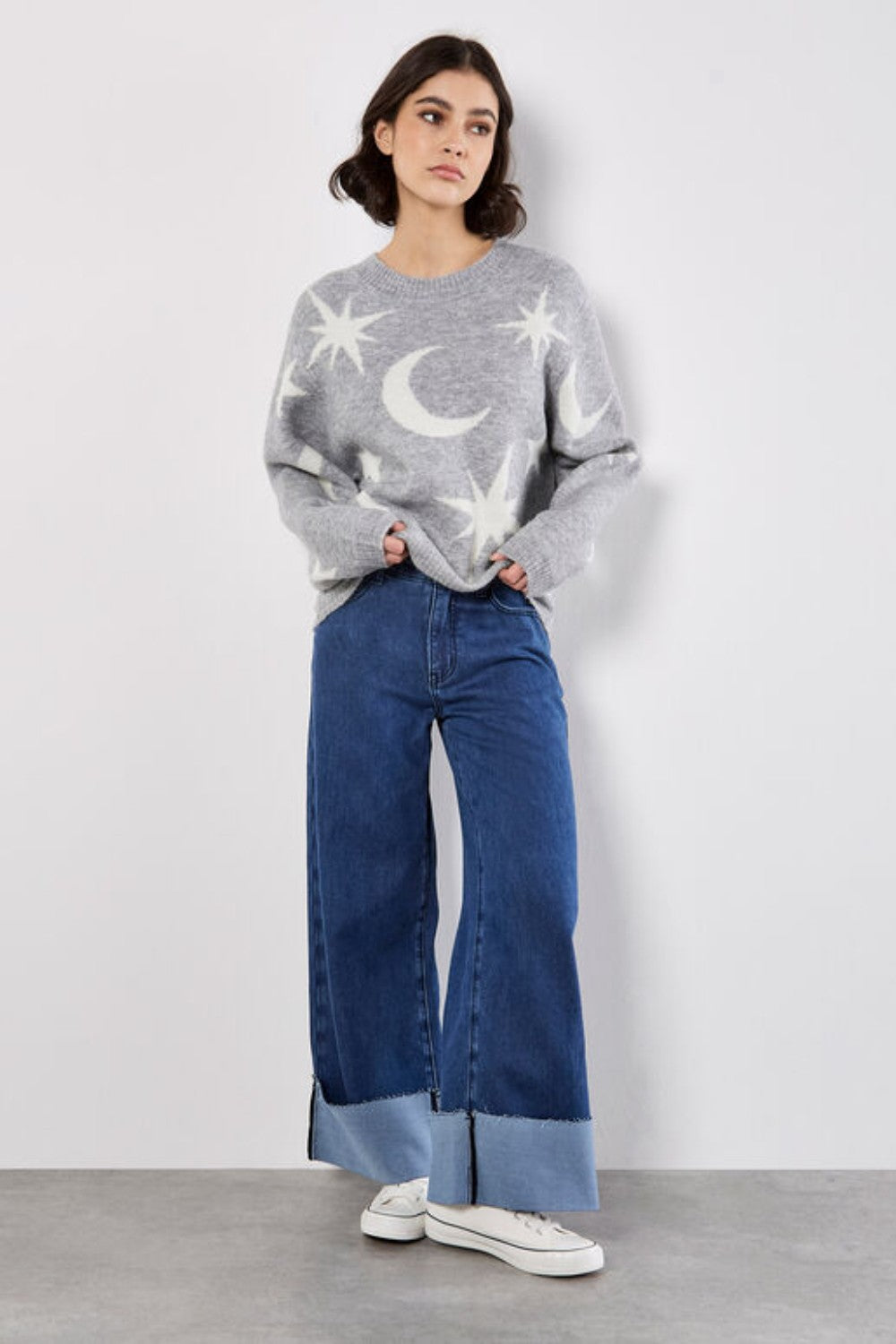Woman wearing a gray sweater with star and moon patterns and blue jeans on a white background