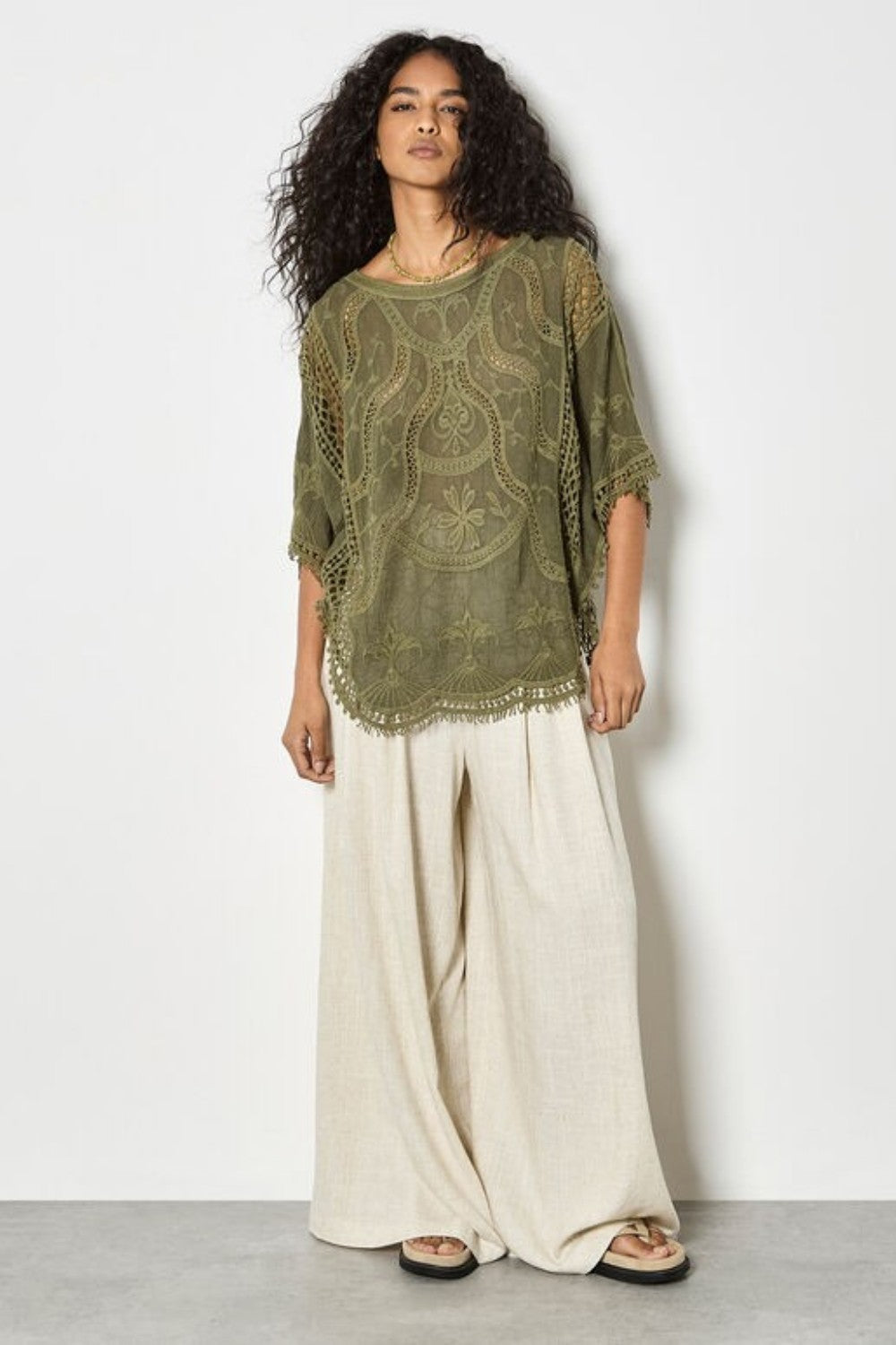 Woman wearing a green lace top and white pants against a light gray background