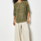 Woman wearing a green lace top and white pants against a light gray background
