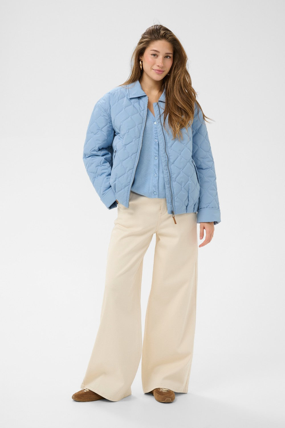 Woman wearing a light blue puffer jacket over a shirt with beige pants on a white background