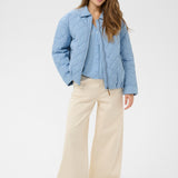 Woman wearing a light blue puffer jacket over a shirt with beige pants on a white background