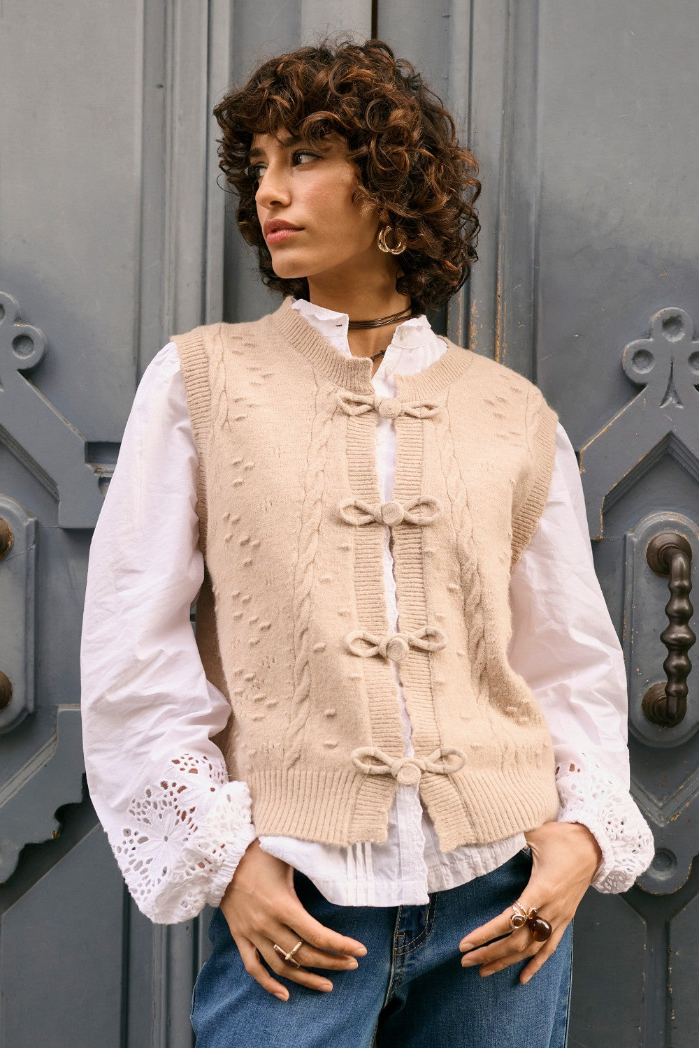 Woman wearing a beige knitted vest over a white blouse with blue jeans, standing against a gray door.