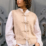 Woman wearing a beige knitted vest over a white blouse with blue jeans, standing against a gray door.