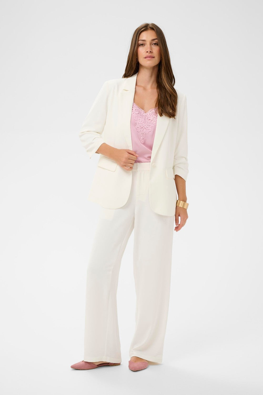Woman wearing a white blazer over a pink top and white pants on a light gray background
