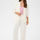 Woman wearing a white blazer over a pink top and white pants on a light gray background