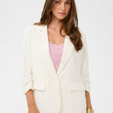 Woman wearing a white blazer over a pink top and white pants on a light gray background