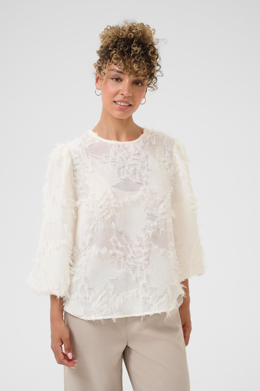 Woman wearing a white lace top and beige pants against a plain background