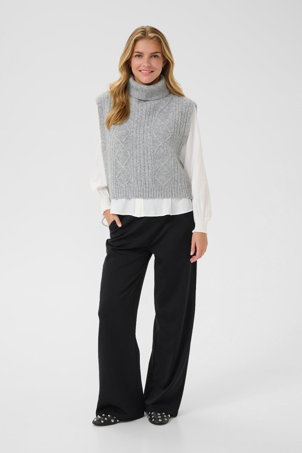 Woman wearing a gray sleeveless sweater over a white shirt with black pants on a white background
