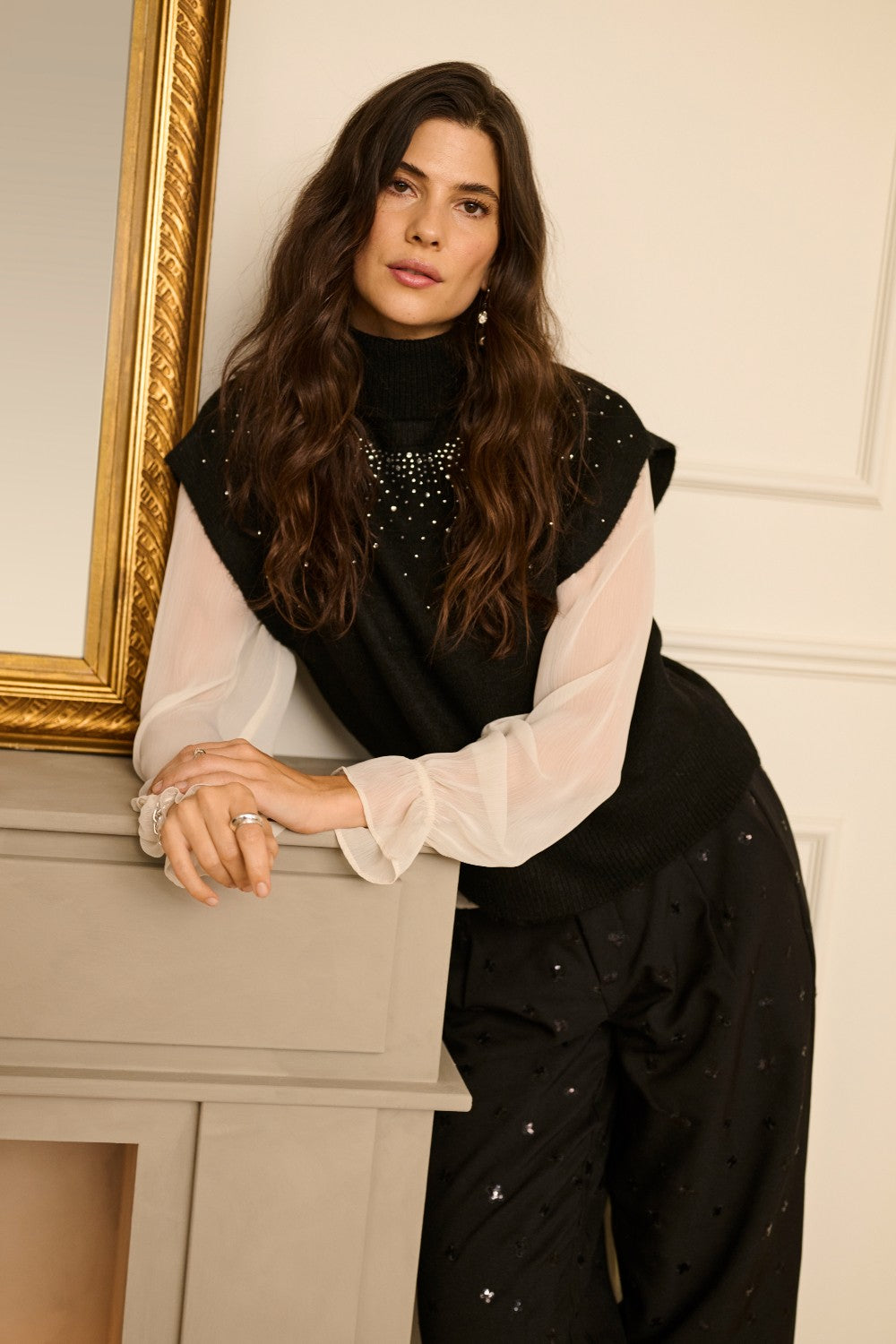 Woman in a stylish outfit with a black top and white sleeves leaning against a fireplace.