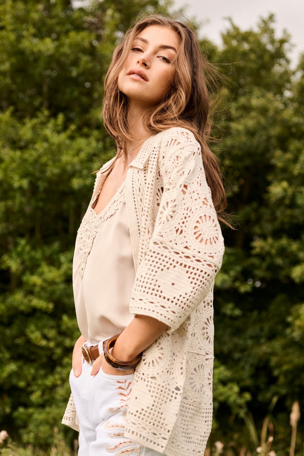 Woman wearing a beige blouse and white pants with a crochet cover-up in a natural setting