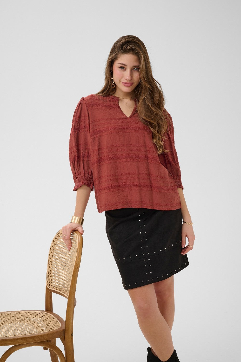 Woman wearing a rust-colored blouse and black skirt standing next to a wooden chair on a white background