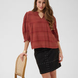 Woman wearing a rust-colored blouse and black skirt standing next to a wooden chair on a white background