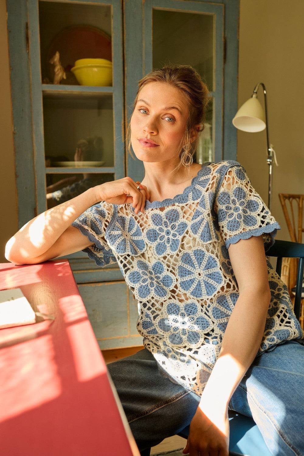 Woman sitting at a table in a room with a lamp and shelves in the background