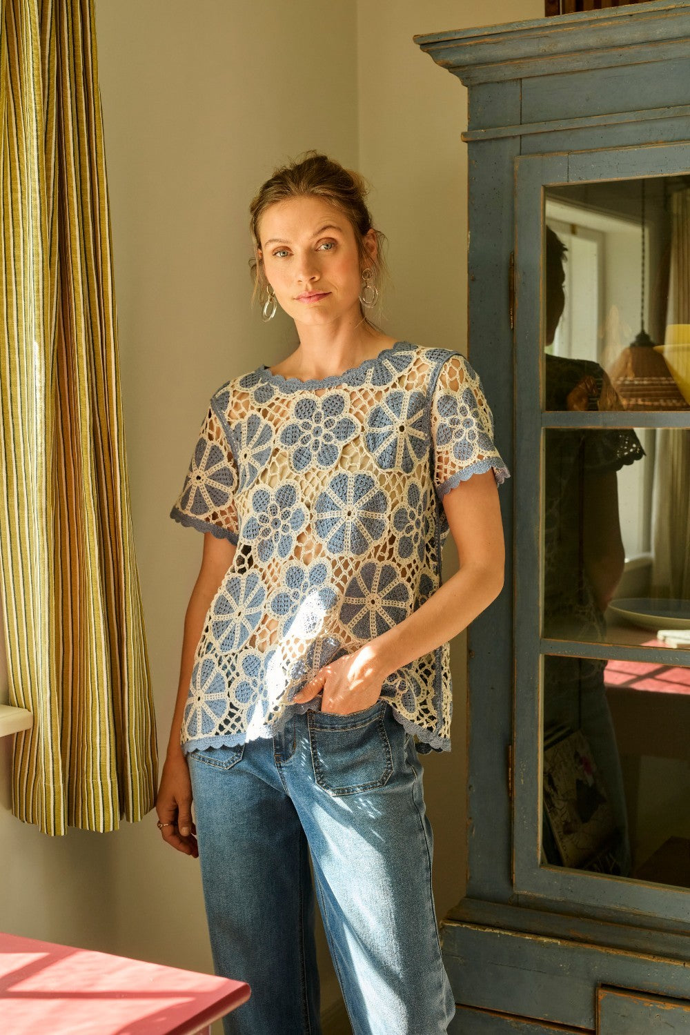 Woman wearing a floral crochet top and jeans standing in a room with a cabinet and curtains.