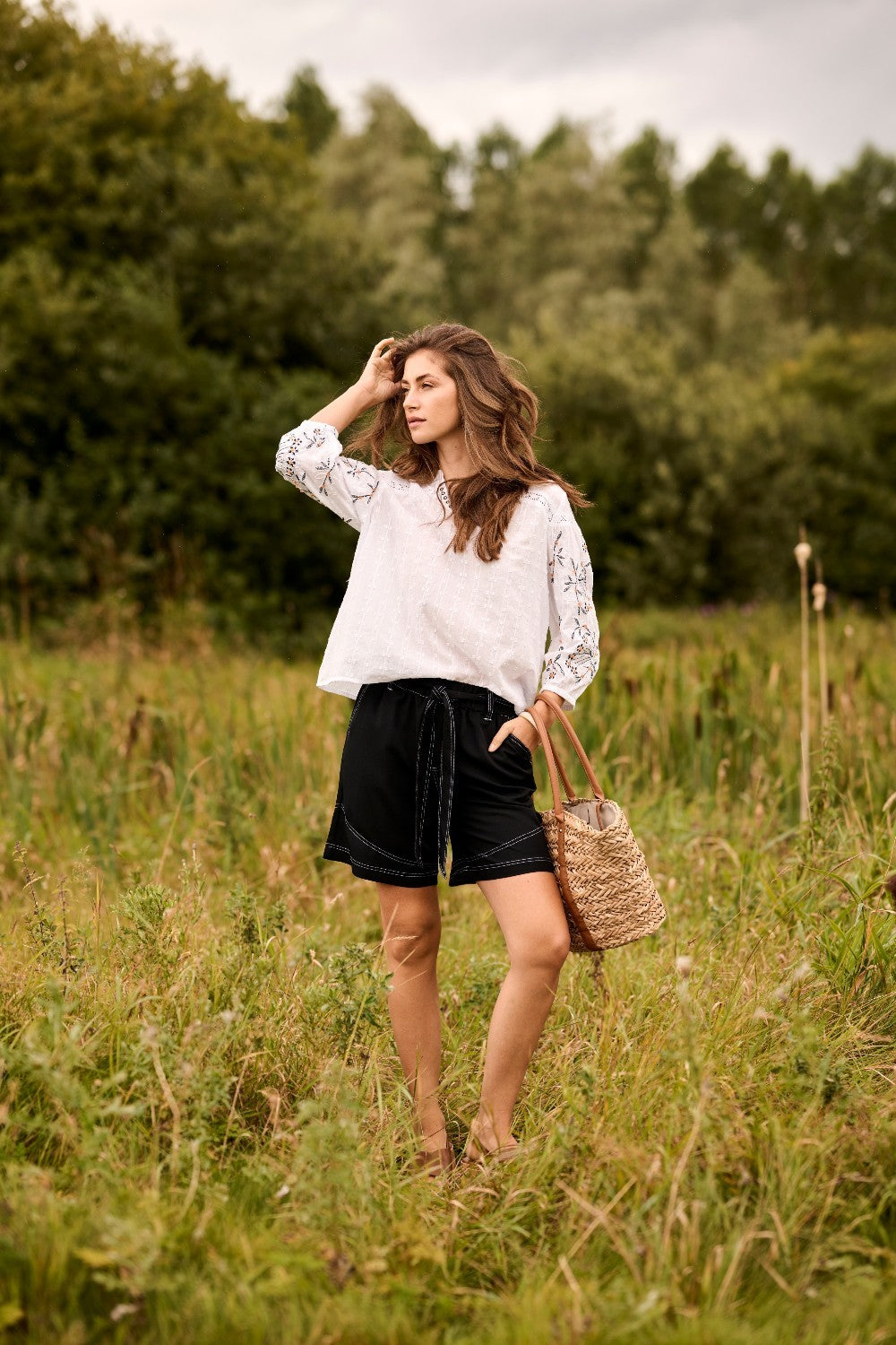 Woman in a white blouse and black shorts standing in a grassy field with trees in the background.