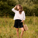 Woman in a white blouse and black shorts standing in a grassy field with trees in the background.