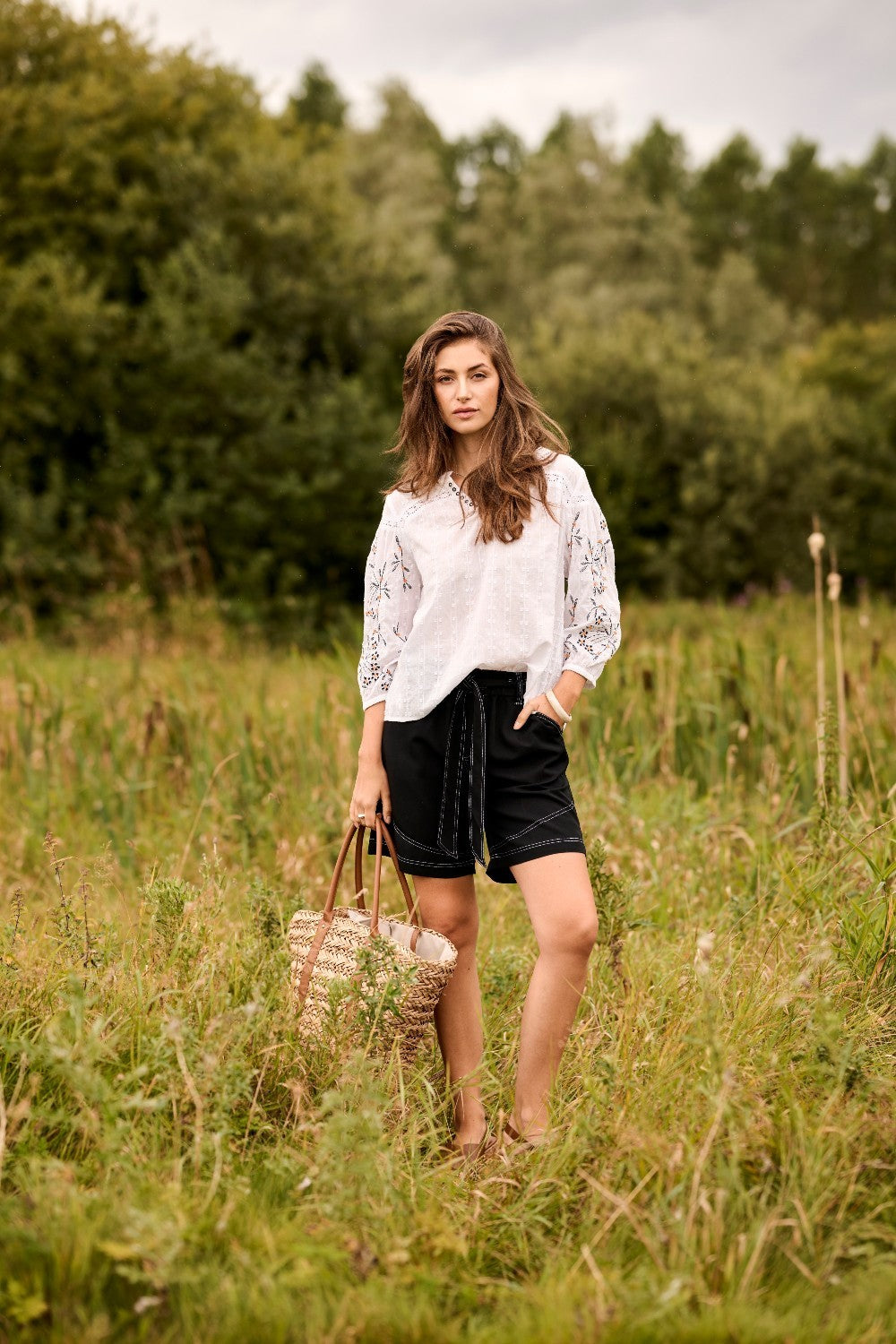 Woman in a white blouse and black shorts standing in a grassy field holding a woven basket.
