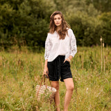 Woman in a white blouse and black shorts standing in a grassy field holding a woven basket.