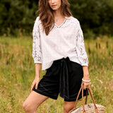 Woman in a white embroidered blouse and black shorts standing in a grassy field.