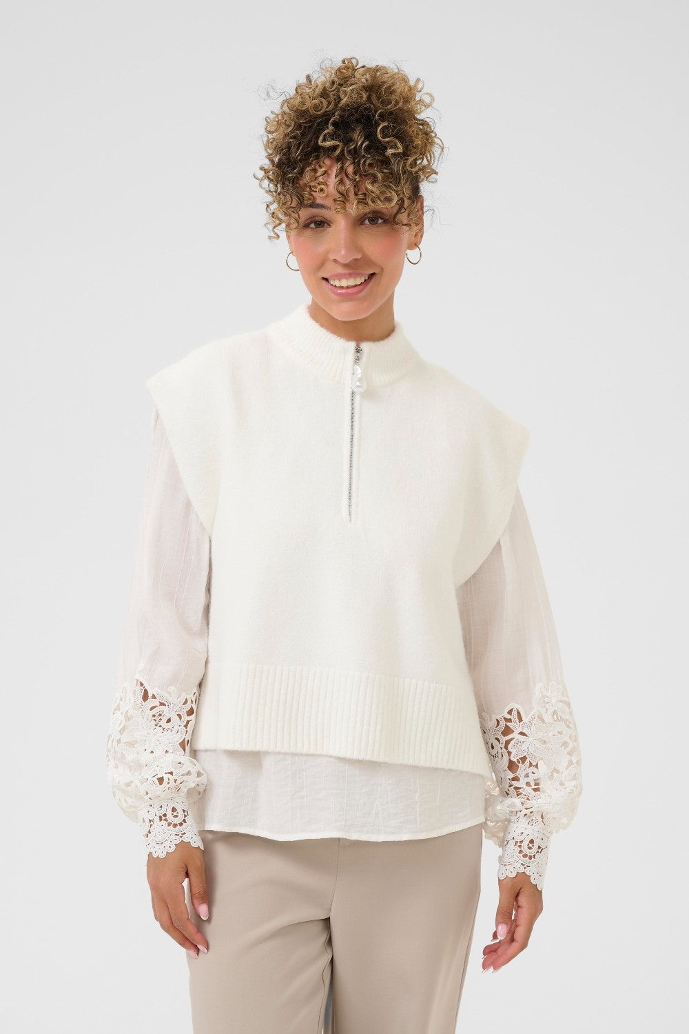 Woman wearing a white vest and blouse with lace sleeves and trim on a plain background